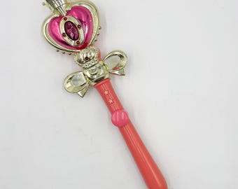 Könnte beinhalten: Ein rosa-goldener Spielzeugstab mit herzförmigem Oberteil, Krone und rosa Griff. Der Zauberstab hat goldene Schleifen- und Sterndetails. Die Oberseite zeigt ein rosa Herz mit einem zentralen Juwel.