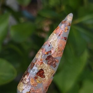 Puede incluir: Primer plano de un esmalte de u&ntilde;as transparente con escamas doradas y cobrizas. El esmalte de u&ntilde;as est&aacute; en un palo blanco con la palabra "Glaze" escrita en &eacute;l.