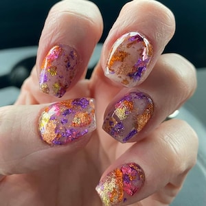 May include: Close-up of five fingernails with a clear base and a design of gold, orange, and purple foil flakes.