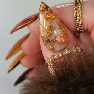 Puede incluir: Primer plano de un dedo con una u&ntilde;a larga, puntiaguda y transparente con detalles de l&aacute;mina dorada y cobre. La u&ntilde;a est&aacute; decorada con un dise&ntilde;o de m&aacute;rmol. El dedo tambi&eacute;n lleva un anillo de oro con peque&ntilde;os diamantes.