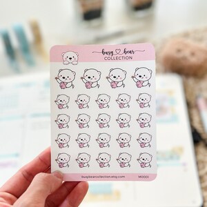 May include: A sheet of stickers featuring a white bear holding a pink piggy bank. The stickers are arranged in a grid pattern on a white background. The text "busy bear collection" and "busybearcollection.etsy.com" are printed at the bottom of the sheet. The sticker sheet is labeled "M0001".