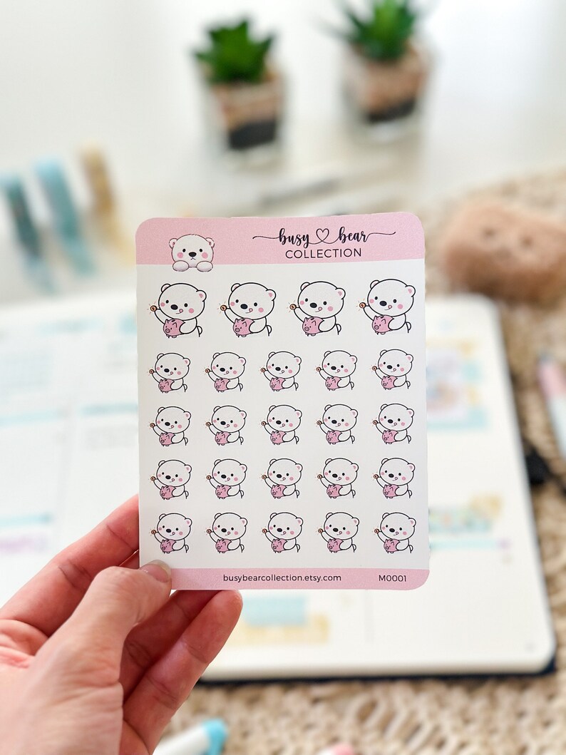 May include: A sheet of stickers featuring a white bear holding a pink piggy bank. The stickers are arranged in a grid pattern on a white background. The text "busy bear collection" and "busybearcollection.etsy.com" are printed at the bottom of the sheet. The sticker sheet is labeled "M0001".