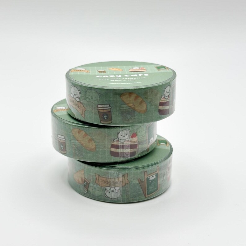 May include: Three rolls of green washi tape stacked on top of each other. The tape is decorated with a repeating pattern of coffee cups, bread, and bear characters. The top of the top roll reads "Cozy Cafe".