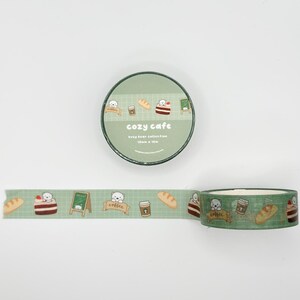 May include: A roll of decorative washi tape with a repeating pattern of a bear, bread, coffee, and cake on a light green background. The tape roll has the words "cozy cafe" printed on it.