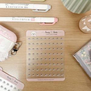 May include: A collection of stationery items on a wooden surface. Includes white Mildliner pens, a pink and white AirPod case, sticker sheets with a bear design, and a notebook. The sticker sheet has the text "busy bear COLLECTION".