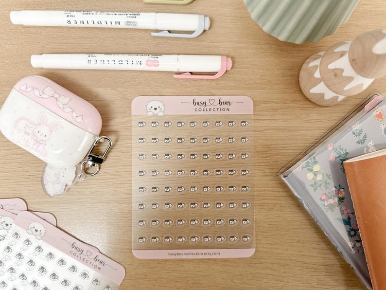 May include: A collection of stationery items on a wooden surface. Includes white Mildliner pens, a pink and white AirPod case, sticker sheets with a bear design, and a notebook. The sticker sheet has the text "busy bear COLLECTION".