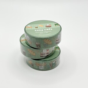 May include: Three rolls of green washi tape stacked on top of each other. The tape features a repeating pattern of bears, coffee cups, and bread. The top roll reads "cozy cafe" in white text.