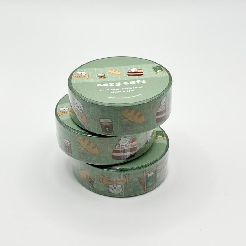 May include: Three rolls of green washi tape stacked on top of each other. The tape features a repeating pattern of bears, coffee cups, and bread. The top roll reads "cozy cafe" in white text.