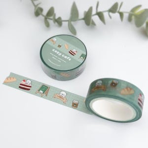 May include: Two rolls of washi tape with a "cozy cafe" theme. The tape is green with illustrations of bears, coffee cups, bread, and cake. One roll is unrolled, showing the repeating pattern. The text "cozy cafe" is visible on the roll.