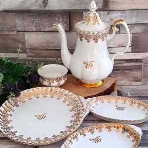 May include: A white teapot with gold trim and the words "Congratulations 50" on the side. The teapot is surrounded by three white plates with gold trim and the same inscription. The plates are on a wooden surface.