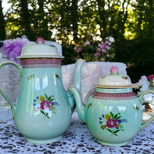 Op de afbeelding: Twee porseleinen theepotten met bloemenmotieven. De grotere theepot is lichtblauw met een roze roos en andere bloemen. De kleinere theepot is lichtblauw met een roze roos en andere bloemen. Beide theepotten hebben gouden accenten.