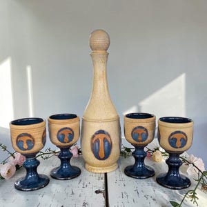 Può includere: Un decanter in ceramica con tappo sferico e quattro calici abbinati con stelo. Ogni pezzo presenta una finitura beige maculata con una base blu scuro e un emblema circolare centrale che mostra una forma arancione stilizzata.