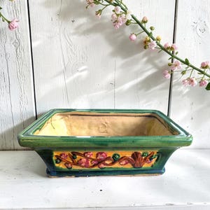 Puede incluir: Una maceta rectangular de cerámica con un borde verde brillante y una banda central amarilla. El exterior presenta un diseño en relieve de ramas marrones con flores rosas y rojas. El interior del recipiente es de un color beige claro y liso.