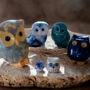 Puede incluir: Una colección de seis pequeñas figuritas de búho de cerámica. Los búhos están pintados en varios tonos de azul, blanco y amarillo. Los búhos están dispuestos sobre una superficie de color marrón claro y texturada.