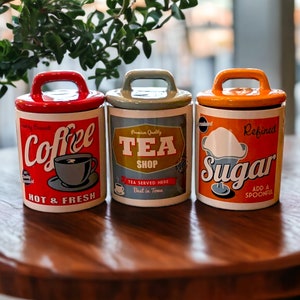 Puede incluir: Tres botes de cerámica con tapas, etiquetados como "Coffee", "Tea Shop" y "Sugar". El bote de café es rojo, el de té es gris y el de azúcar es naranja. Cada uno tiene un asa y etiquetas de estilo vintage.