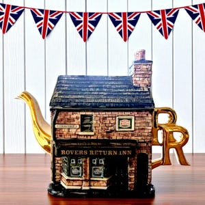 Puede incluir: Tetera decorativa con forma de edificio de ladrillo, con boquilla y asa doradas. El diseño del edificio incluye el texto "ROVERS RETURN INN" y "CORONATION ST". Guirnaldas de banderas de la Union Jack cuelgan encima.