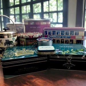 Puede incluir: Surtido de cajas de hojalata vintage con varios diseños. Las cajas presentan un diseño verde y dorado con una corona, un diseño floral verde y blanco, un diseño rojo y blanco con un autobús y un diseño blanco y dorado con un coche. Las cajas están sobre una superficie de madera.