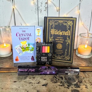 Può includere: Un assortimento di articoli spirituali, tra cui un mazzo di tarocchi, un libro intitolato "Witchcraft" e una scatola di "Magic Spell Candles". Sono presenti anche bastoncini di incenso e candele accese.