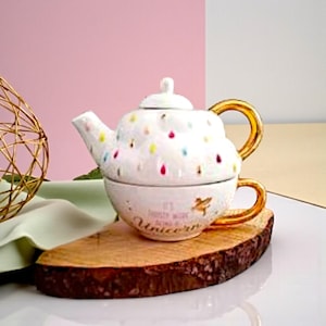 Peut inclure: Un ensemble théière et tasse en céramique blanche avec des anses dorées. La théière est ornée de pois colorés et d'un couvercle. La tasse porte l'inscription "It's frosty work being a Unicorn" et un motif de licorne dorée. L'ensemble est posé sur une tranche de bois.