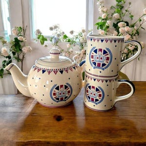 May include: A white teapot with a blue and red crown design and a matching set of three mugs. The teapot and mugs have a white background with red and blue polka dots and a red, white, and blue bunting pattern.