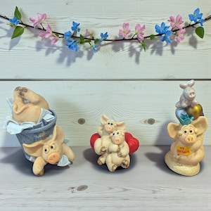 Może przedstawiać: Kolekcja fantazyjnych figurek świń. Jedna świnia jest w koszu na śmieci, inna obejmuje dwa prosięta, a trzecia trzyma znak z napisem "My Birthday". Figurki są beżowe z wyrazistymi twarzami. Girlanda z różowych i niebieskich kwiatów zdobi tło.