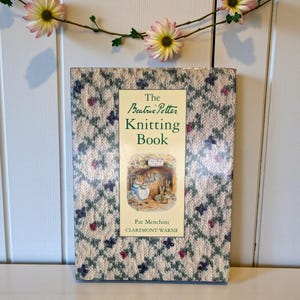 May include: A hardcover book titled "The Beatrix Potter Knitting Book" with a patterned cover in cream, blue, and burgundy. The book's cover features a central illustration and text. A floral garland hangs above the book.
