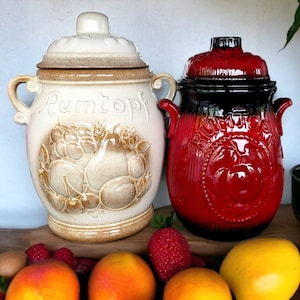 May include: Two ceramic jars with lids, one white with brown accents and the word "Rumtopf" on the front, the other red with black accents. Both jars have handles. The jars are sitting on a wooden surface with fruit and other items around them.