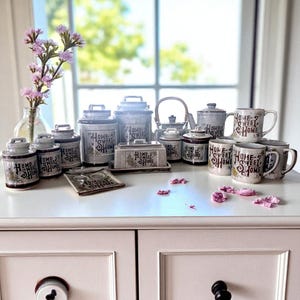 Peut inclure: Une collection d'articles de cuisine de style vintage, comprenant des boîtes, des tasses, une théière et un beurrier, disposés sur une surface blanche. Les articles sont principalement blancs et gris, avec l'inscription "HOME SWEET HOME". Un vase de fleurs roses est en arrière-plan.