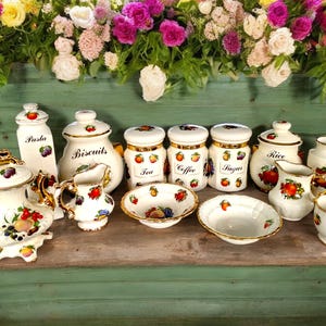 Puede incluir: Un juego de botes de cocina de cerámica blanca con ribetes dorados e ilustraciones de frutas. Los botes están etiquetados como "Pasta", "Biscuits", "Té", "Café", "Azúcar" y "Arroz". También hay una tetera, una jarra de crema, un azucarero y una taza.