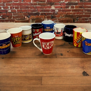 Puede incluir: Una colección de 10 tazas de cerámica con varios logotipos de barras de chocolate, incluyendo Yorkie, Toffee Crisp, Twirl, Maltesers, KitKat, Time Out, Smarties y una segunda taza Yorkie. Las tazas están sobre una mesa de madera.
