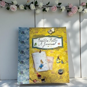 May include: A hardback journal with a yellow cover, featuring the text "Beatrix Potter A Journal". The journal has a blue floral patterned spine and illustrations of Peter Rabbit. A butterfly and other decorative elements are also present.