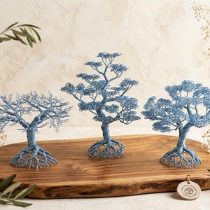 May include: Three decorative wire trees crafted from light blue metallic wire, each featuring a twisted trunk, exposed root system, and a dense canopy of looped wire branches. The trees are arranged on a smooth, oval-shaped wooden board.