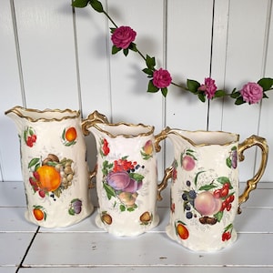 Pode incluir: Três jarras de cerâmica creme com acabamentos e alças douradas. Cada jarra é decorada com ilustrações coloridas de frutas, incluindo laranjas, uvas, ameixas e bagas. As jarras estão dispostas sobre uma superfície branca com uma grinalda floral no fundo.
