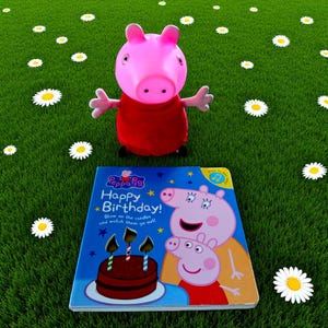May include: A pink Peppa Pig toy figure in a red dress is positioned near a children's book titled "Happy Birthday!". The book cover shows Peppa Pig and a birthday cake with candles. The scene is set on a green lawn with scattered daisies.