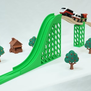 May include: A toy train set featuring a tall green plastic ramp and bridge supports, wooden tracks, a white and red train engine with a green cargo car, a brown miniature house, and several small green trees with red fruit accents.