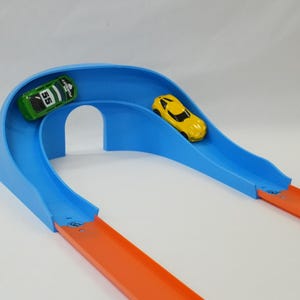 May include: A blue toy race track with a curved design, featuring a green and a yellow toy car. The track has orange lanes and a bridge-like structure. A fun toy for children.