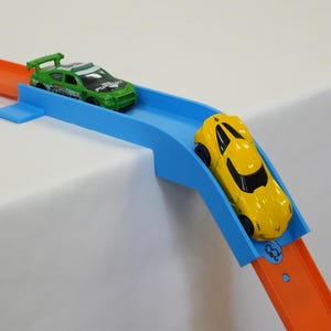 May include: A blue plastic track connector piece joins two orange toy car tracks. A green toy race car with a rear spoiler sits on the flat section, while a yellow toy sports car is positioned on the curved downward slope of the blue connector.