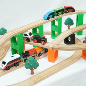 May include: A wooden toy train set featuring multiple tracks elevated by green and orange block supports. The set includes various colorful toy trains, small plastic trees with red fruit, and a brown wooden house structure on the tracks.