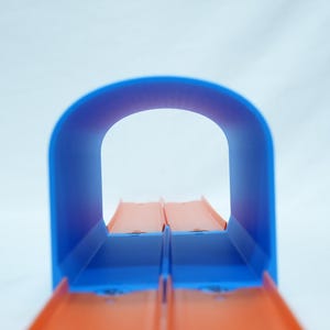 Pu&ograve; includere: Un tunnel per pista giocattolo blu e arancione. Il tunnel ha una parte superiore curva e due piste arancioni parallele. Lo sfondo &egrave; di colore bianco.