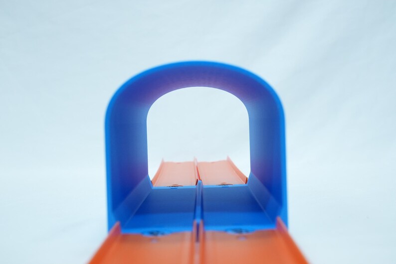 Pu&ograve; includere: Un tunnel per pista giocattolo blu e arancione. Il tunnel ha una parte superiore curva e due piste arancioni parallele. Lo sfondo &egrave; di colore bianco.