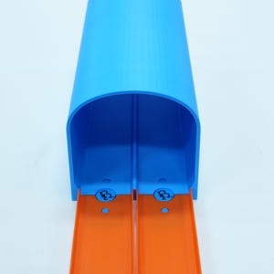 Pu&ograve; includere: Un tunnel di pista giocattolo in plastica blu con due corsie arancioni. Il tunnel ha una parte superiore arrotondata ed &egrave; progettato per le macchinine. La pista &egrave; su sfondo bianco.