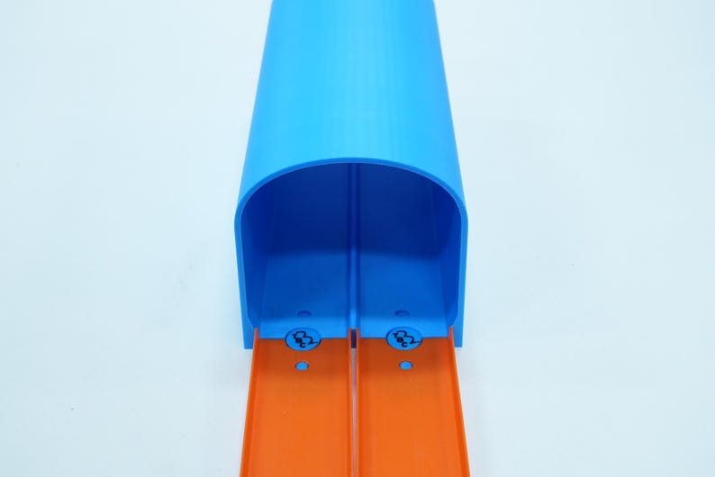 Pu&ograve; includere: Un tunnel di pista giocattolo in plastica blu con due corsie arancioni. Il tunnel ha una parte superiore arrotondata ed &egrave; progettato per le macchinine. La pista &egrave; su sfondo bianco.