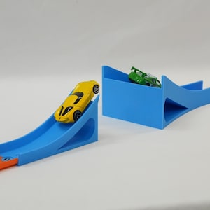 May include: A yellow toy car rolls down a blue ramp, part of a toy race track set. A green toy car sits in a blue structure. The track is orange. The set is designed for toy cars.
