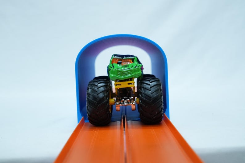 Pu&ograve; includere: Un camion mostro giocattolo verde e nero con grandi pneumatici neri &egrave; posizionato su una pista arancione, diretto verso un arco blu. Il camion giocattolo ha un design dettagliato ed &egrave; pronto per la corsa.