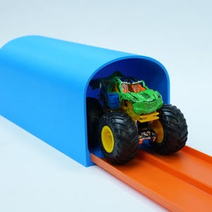 Pu&ograve; includere: Un monster truck giocattolo con grandi pneumatici neri e una carrozzeria verde e blu &egrave; posizionato su una pista arancione, che entra in un tunnel blu brillante. La pista &egrave; progettata per le macchinine ed &egrave; su sfondo bianco.