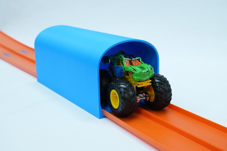 Pu&ograve; includere: Un monster truck giocattolo con grandi pneumatici neri e una carrozzeria verde e blu &egrave; posizionato su una pista arancione, che entra in un tunnel blu brillante. La pista &egrave; progettata per le macchinine ed &egrave; su sfondo bianco.