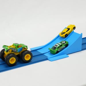 May include: A toy race track with three miniature vehicles: a yellow sports car, a green race car, and a green and yellow monster truck. The track is blue with a ramp for the cars.