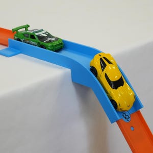 May include: A toy car race track with two miniature cars. One car is green and the other is yellow. The track is blue and orange, set against a white background.