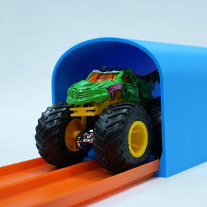 Pu&ograve; includere: Un giocattolo di monster truck verde e giallo con grandi pneumatici neri e cerchi gialli &egrave; posizionato sotto un arco blu. Il camion &egrave; su una pista arancione, suggerendo una gara o un gioco. Lo sfondo &egrave; bianco.