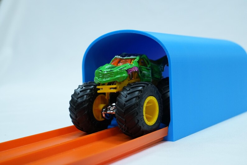 Pu&ograve; includere: Un giocattolo di monster truck verde e giallo con grandi pneumatici neri e cerchi gialli &egrave; posizionato sotto un arco blu. Il camion &egrave; su una pista arancione, suggerendo una gara o un gioco. Lo sfondo &egrave; bianco.